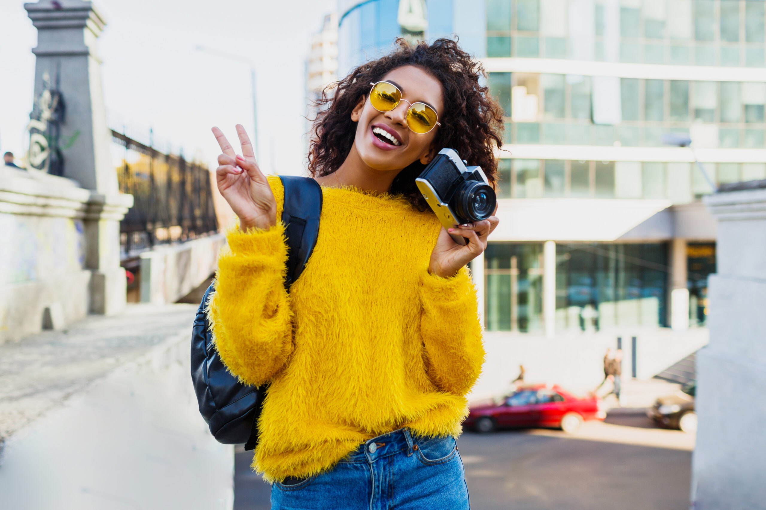 Successful  black female photographer making photos on modern architecture background. Wearing back pack, yellow sweater and glasses.