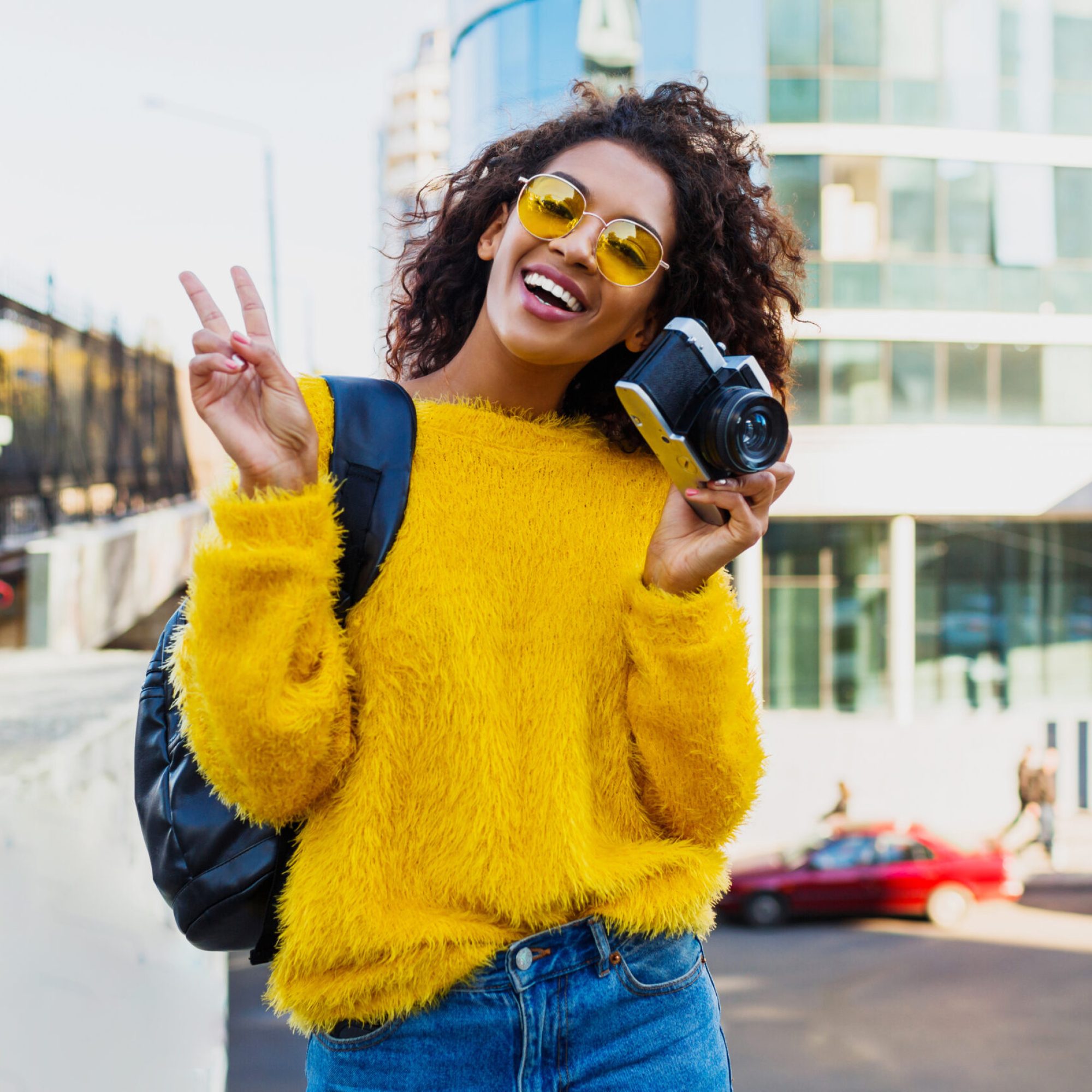 Successful  black female photographer making photos on modern architecture background. Wearing back pack, yellow sweater and glasses.